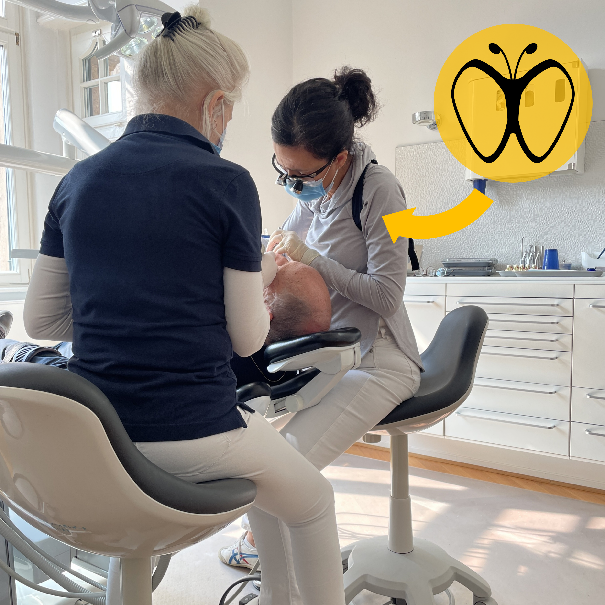 Eine Zahnรคrztin trรคgt den StraightUp Haltungstrainer wรคhrend einer Zahnbehandlung. Sie sitzt konzentriert รผber einem Patienten, wรคhrend eine Assistentin daneben steht. Ein gelber Pfeil hebt das Logo des Haltungstrainers hervor.