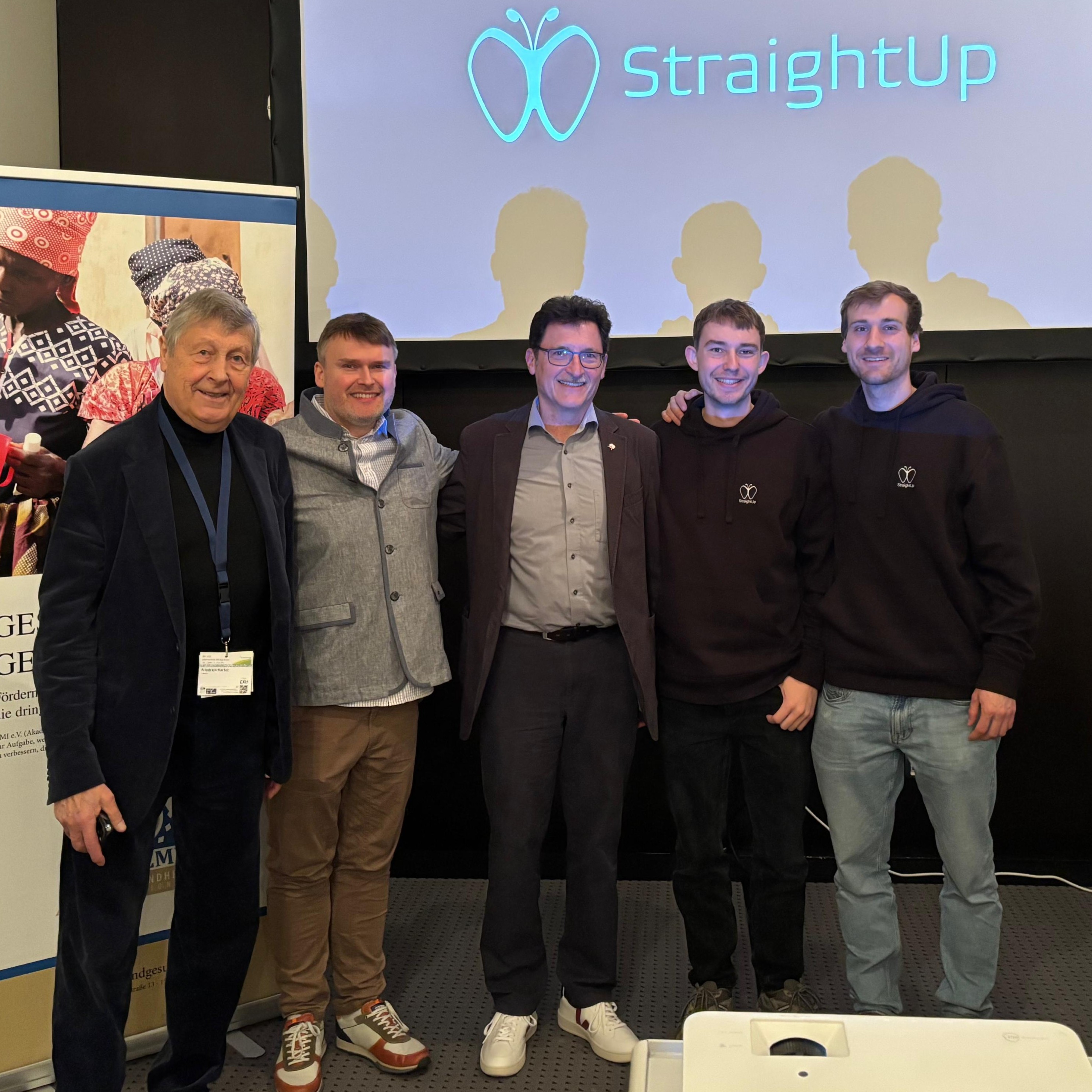 Gruppenfoto nach dem Kolloquium: Zwei Teammitglieder von StraightUp posieren zusammen mit drei weiteren Mรคnnern vor einer Leinwand mit dem StraightUp-Logo. Alle lรคcheln in die Kamera, der Moment wirkt feierlich und gelungen.