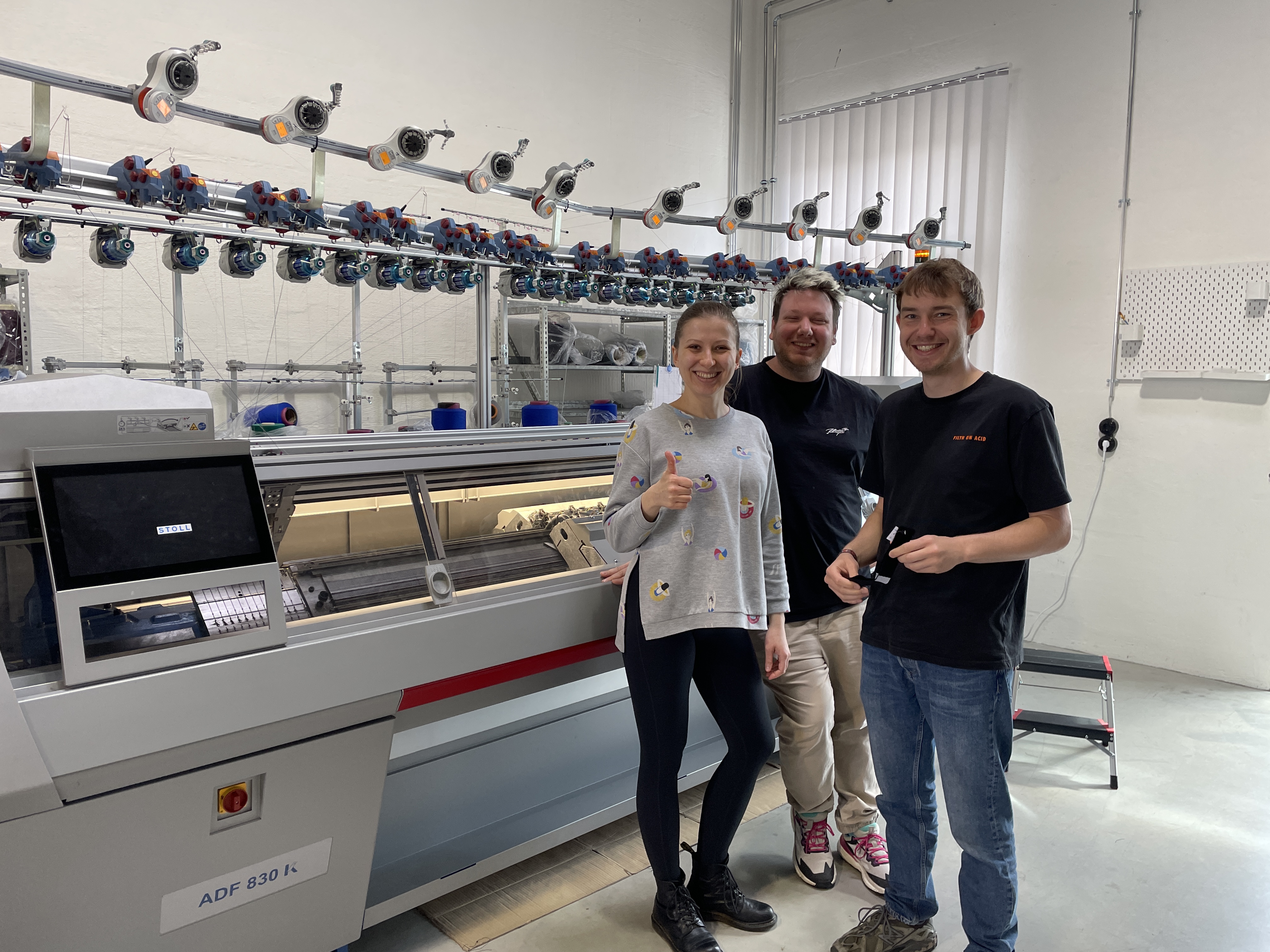 Drei Personen stehen lachend vor einer industriellen Strickmaschine im Smart Textiles Hub. Eine Frau zeigt Daumen hoch, die beiden Mรคnner daneben halten Materialproben in der Hand. Im Hintergrund sind Garnspulen und technische Ausrรผstung sichtbar โ die Szene vermittelt Fortschritt und Zusammenarbeit in der Textilentwicklung.