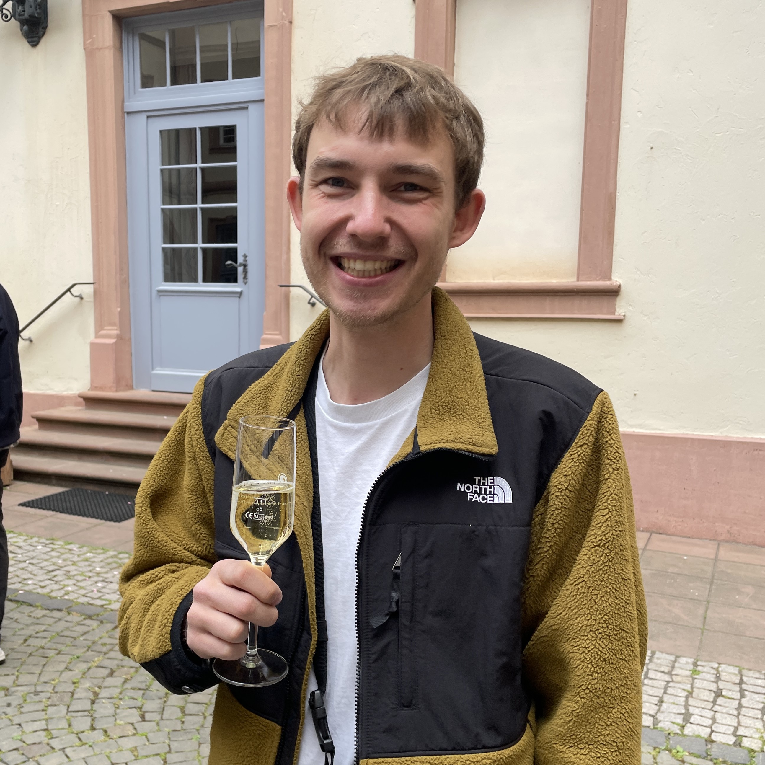 Eine lรคchelnde Person hรคlt ein Glas Sekt in der Hand und steht vor einem historischen Gebรคude mit hellem Putz und rosa Fensterrahmen. Die Stimmung wirkt feierlich und entspannt โ ein Moment zum Anstoรen und Feiern.
